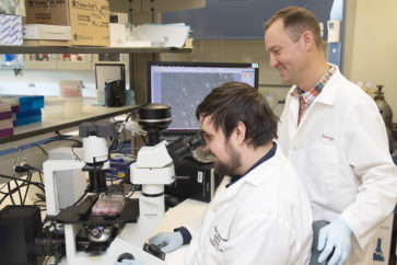 A researcher mentors a research student standing behind him as he looks through a microscope
